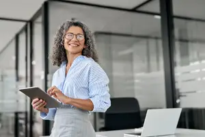 A smiling woman with glasses standing in an office room holding a tablet