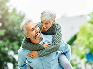 An older man carrying a woman on his shoulders outside in a garden with trees and a house in the background