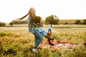 A family of three is playing in a grassy field during the daytime, with one person carrying a child on their back, another child running and holding a football, and a blanket on the ground.