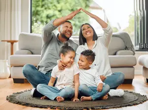 A family of four, consisting of two adults and two children, are sitting on a rug in a living room with a glass window in the background.