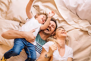 A family of three is lying on a bed with white sheets and pillows, with the child smiling and raising both hands, the man holding the child with a smile, and the woman lying on the bed with a pillow behind her head and a smiling face.