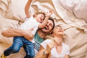 A family of three is lying on a bed with white sheets and pillows, with the child smiling and raising both hands, the man holding the child with a smile, and the woman lying on the bed with a pillow behind her head and a smiling face.