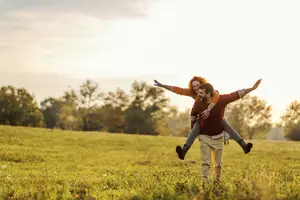 A man and a woman are walking in a grassy field with trees in the distance, and the man is carrying the woman on his back while they are both smiling and looking at the camera. They are both wearing long-sleeved shirts, pants, and boots, and the man is wearing a watch and a ring. The sky is cloudy, and the sun is setting behind them, casting a warm glow on the scene.