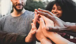 Three people, two women and a man, smiling and putting their hands together in a huddle, possibly in a park during the day.