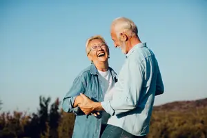An elderly man and woman are smiling and laughing while holding hands in an outdoor area with trees and mountains in the background.