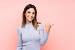 A woman is standing in front of a pink wall and smiling while pointing at something with her right hand