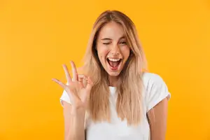 A woman with blonde hair is making a peace sign with her hand and smiling while posing for a photo in front of a yellow background.