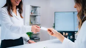 Two women in white lab coats are exchanging a piece of paper at a desk in a clinic.