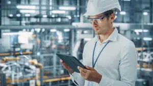A man in a white hard hat and goggles is looking at a tablet in a factory.