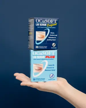 A person holding two boxes of Ocusoft Lid Scrub Plus eye pads, one for mild-moderate conditions and the other for moderate-severe conditions, on a blue background.