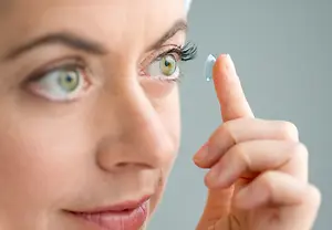A woman wearing a white towel on her head is holding a contact lens in her right hand and touching her eye with her left hand.
