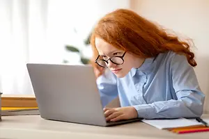 A young girl with glasses and red hair is typing on a laptop on a desk.