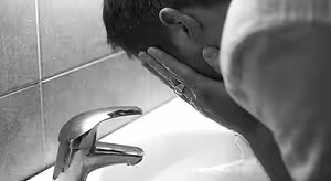A man washes his face at a sink in a bathroom.