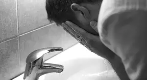 A man washes his face at a sink in a bathroom.