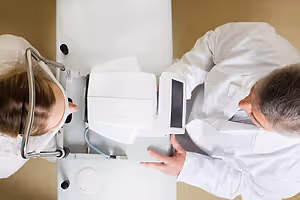 A man and a woman, both wearing white coats, are in a medical examination room, using an ophthalmoscope to examine the woman's eyes.