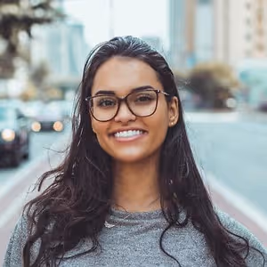 A woman with glasses and a necklace smiles on a city street