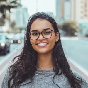 A woman with glasses and a necklace smiles on a city street