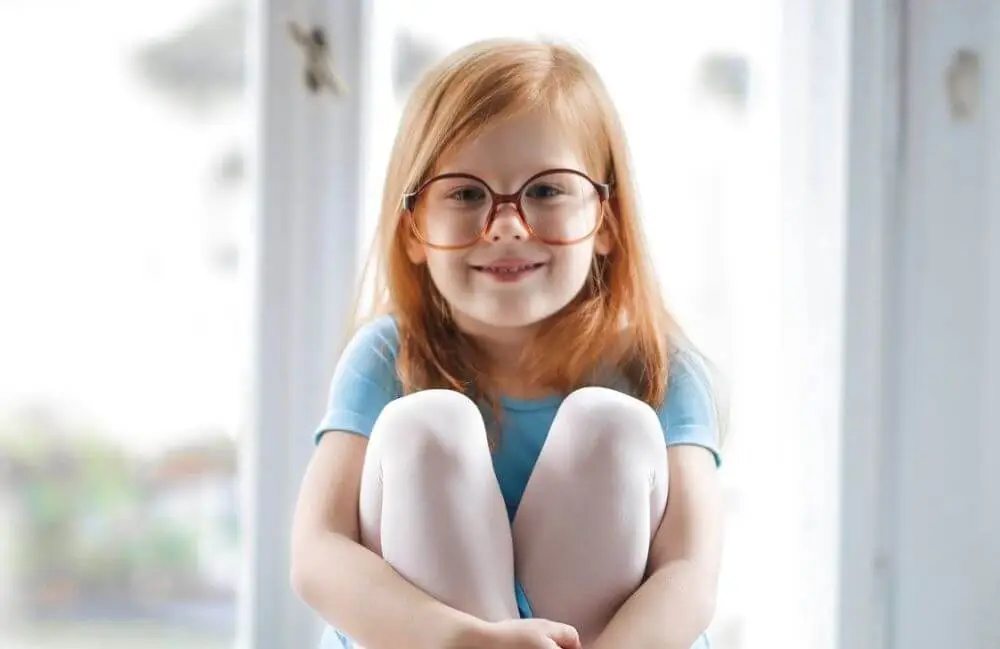 A little girl with glasses and a blue shirt is sitting on the floor in front of a glass window.