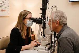 A woman wearing a face mask is examining a man's eyes with a medical device.