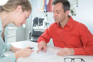 An adult male is sitting at a desk with a woman, both of them are reading a piece of paper.