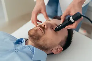 A man lying on a table while a doctor holds a black medical tool against his face.