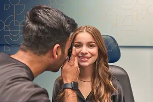 A man wearing a gray shirt and watch is examining a woman's eyes with an ophthalmoscope in a blue chair