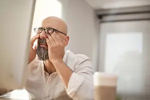 A bald man with a beard wearing glasses is covering his eyes with his hand while sitting in front of a computer monitor