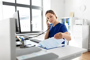 A woman in blue scrubs is speaking on the phone while looking at papers on her desk.