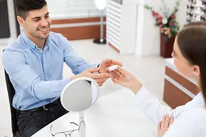 A man and a woman are sitting in a room, the woman is examining the man's eyeglass and the man is smiling.