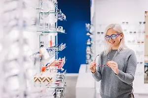 A woman wearing glasses and a gray long-sleeve sweater is smiling and looking at glasses on a shelf in an optical shop.