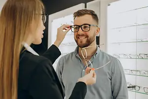 A man is adjusting his glasses with a woman standing next to him in a store with eyeglasses on display