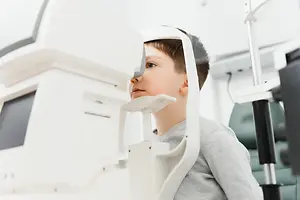 A young boy wearing a gray sweater is looking through a white eyeglass machine in an indoor area.