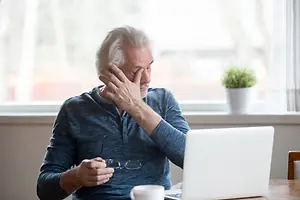 An older man sitting in a chair in front of a laptop with his hand on his forehead and a plant in the window behind him.