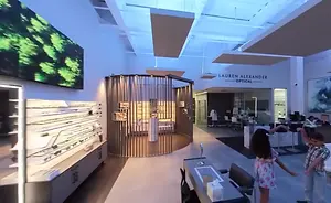 The interior of the Lauren Alexander Optical shop, featuring a modern design with a large monitor on the wall, a spiral staircase, and children playing in the foreground.