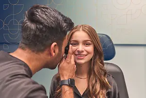 A young woman smiles as a male optometrist examines her eyes with an ophthalmoscope
