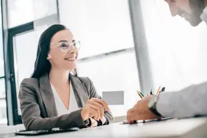 A smiling woman wearing glasses is handing a card to a man in an office setting.