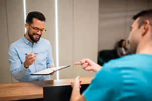 A man wearing glasses is smiling and holding a pen and clipboard in his hand while talking to another man in a blue shirt in a room