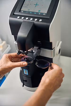 A person wearing a ring is holding a pair of glasses in front of a machine with a digital screen, probably in a laboratory.