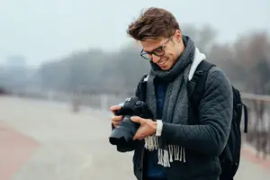 A man is holding a camera and is smiling while wearing a jacket, scarf, and glasses. He is carrying a backpack on his back. Behind him is a blurry background of a bridge and trees. The sky is gray and has some clouds. The man seems to be taking a picture.