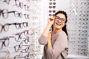 A woman is smiling and posing with a pair of glasses on a rack of glasses in a store.