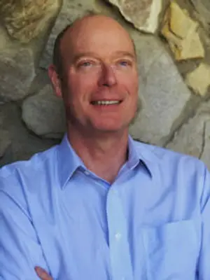 A man with a bald head wearing a blue shirt is smiling and posing for a photo in front of a stone wall.