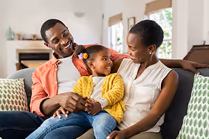 A family of three people sitting on a couch in a living room. The father is holding his daughter's hands, and the mother is smiling while looking at the camera. They are wearing casual clothing, and the couch has pillows with a leaf pattern. Behind them, there is a fireplace and a glass window with blinds. 