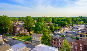 A cityscape view of Lancaster, Pennsylvania, featuring a mix of residential houses, commercial buildings, and lush green trees under a blue sky with scattered clouds, highlighting the vibrant urban landscape.