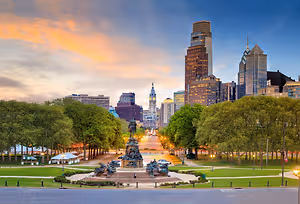 A cityscape featuring a prominent statue at the center, surrounded by trees and buildings under a blue sky