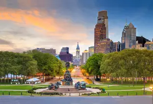 A cityscape featuring a prominent statue at the center, surrounded by trees and buildings under a blue sky