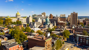 An aerial view of a city with a mix of modern and historic buildings, lush green trees, and a clear blue sky with a few clouds.