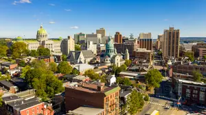 An aerial view of a city with a mix of modern and historic buildings, lush green trees, and a clear blue sky with a few clouds.