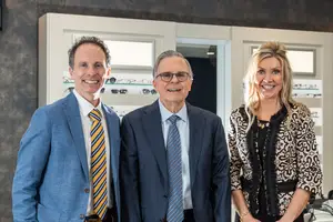 Three people in formal attire stand together smiling in a room with a shelf of glasses in the background