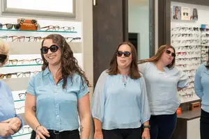 Four women are standing in a shop while smiling, one of them is wearing a necklace, and they are all wearing sunglasses and blue blouses. Behind them are several glasses on display on shelves. One of the women is looking to the left, while another is looking to the right, and the other two are looking at something. A mirror is on the wall behind them.