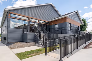 Image of a modern house with gray exterior walls and black railings, featuring a glass door and windows, under a blue sky with some clouds, with a walkway and stairs leading up to the entrance